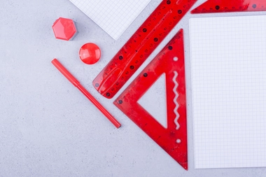 Red rulers, protractor, pen, and geometric shapes on a grey surface with graph paper in the background.